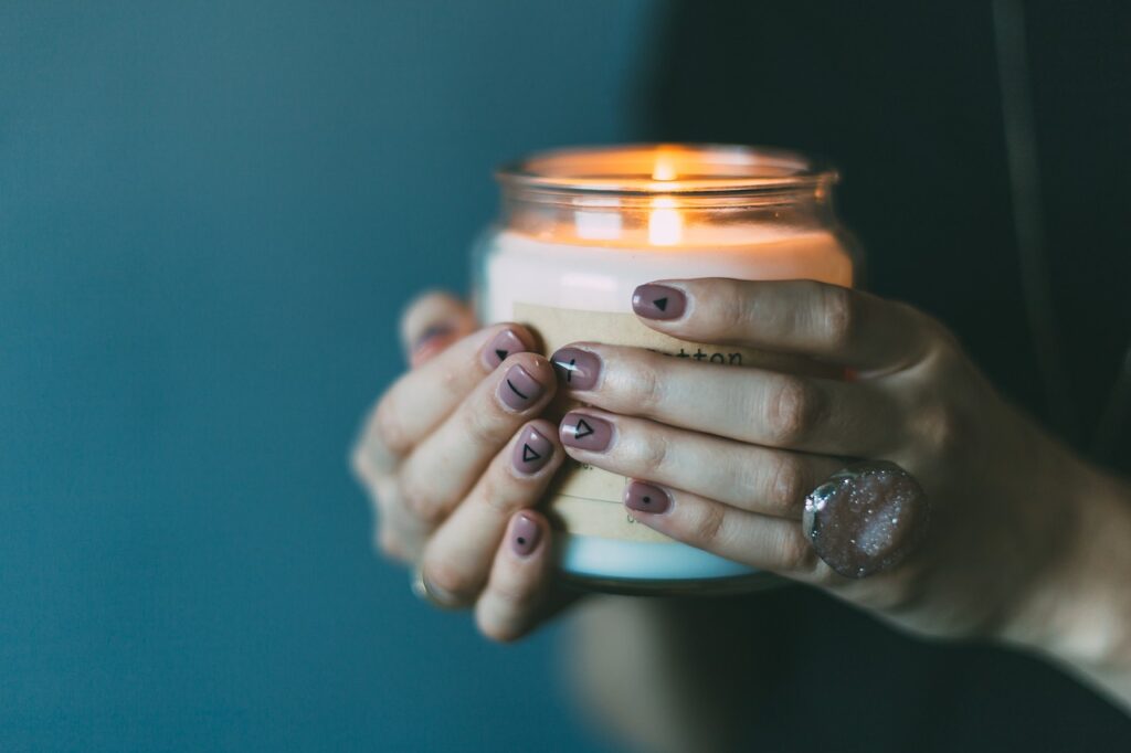 A ladies hands with symbols on her fingernails, holding a lit candle in a jar.